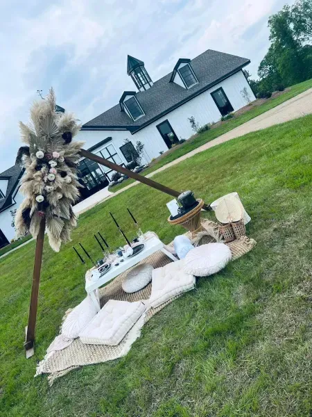 Boho picnic setup on grass with pillows, rug, candles, and pampas grass near a white house