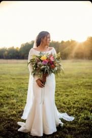 Person in a white dress holding a bouquet in a grassy field at sunset