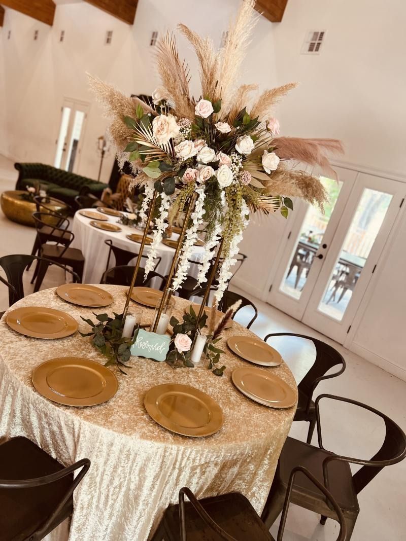 Decorated round banquet table with gold chargers and tall floral centerpiece in a bright event hall