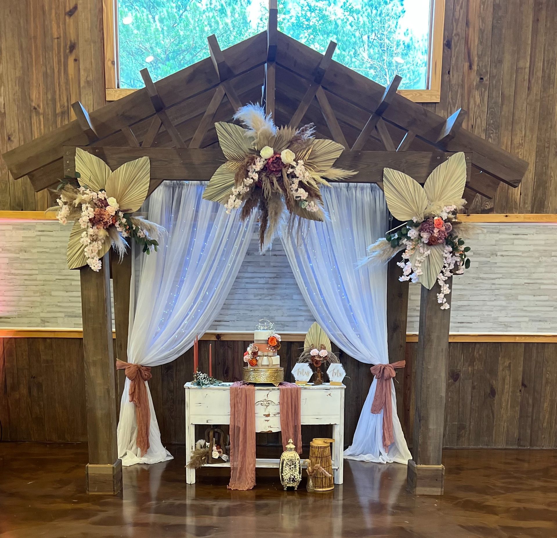 Rustic chapel altar with white drapes, floral arrangements, and candles against wood-paneled walls.
