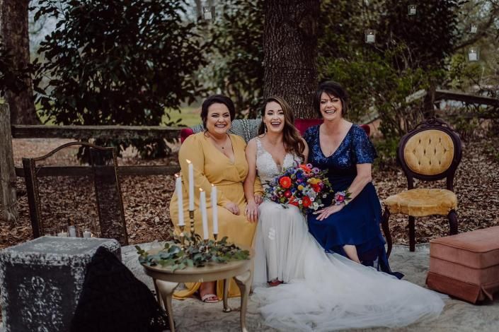 Three women posing outdoors at a wedding, with a bride holding a colorful bouquet.