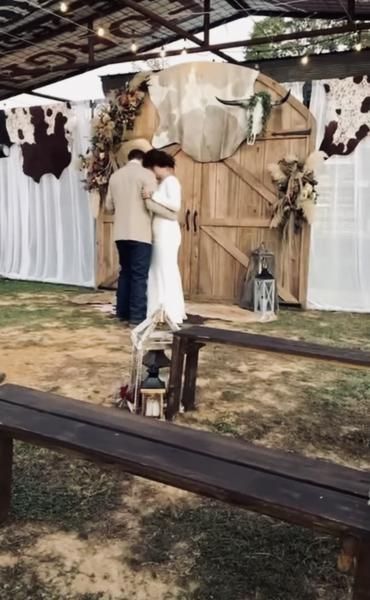 Two people stand by a rustic wooden booth with white drapes and hanging decorations in an outdoor setting.