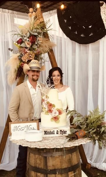 Couple posing behind a decorated barrel wedding cake table with floral arch and white drapery