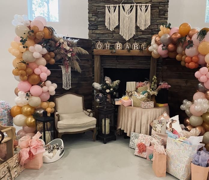 Boho baby shower backdrop with pastel balloon arches, fireplace decor, gifts, and a cream chair