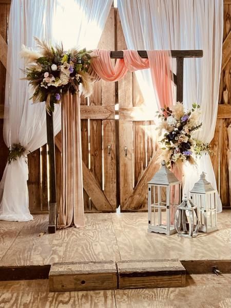 Rustic wedding arch with white drapes, pink fabric, floral arrangements, and lanterns on a wooden stage