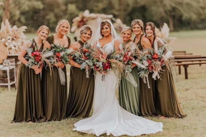 Bride with six bridesmaids in olive dresses holding bouquets outdoors at a wedding venue
