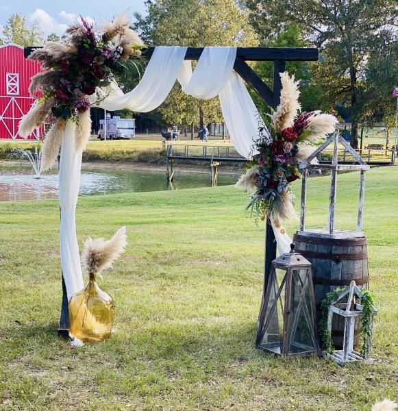 Outdoor wedding arch draped in white fabric and flowers beside a pond, with lanterns and a gold-clad decorative figure