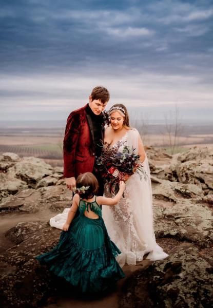 Three people in formal attire standing on rocky terrain under a cloudy sky, one in a red jacket and two in dresses.