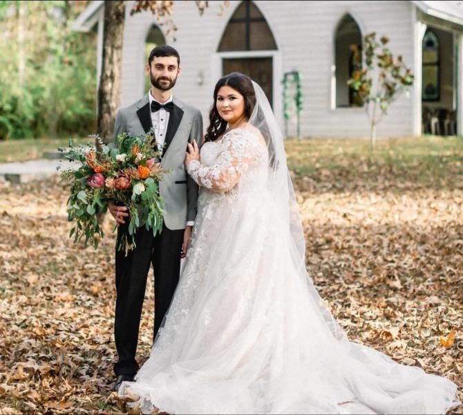 Newlywed couple posing outdoors beside a church, holding a bouquet, with autumn leaves and a long white veil