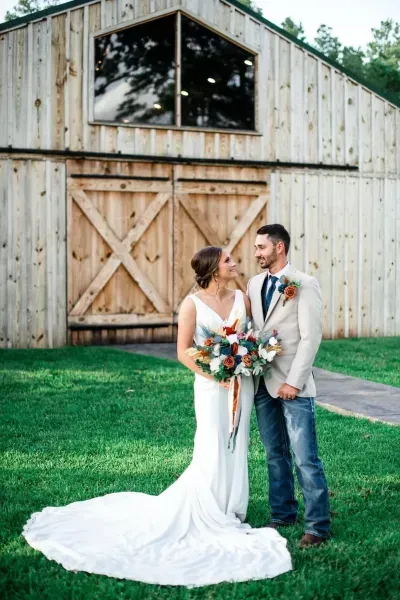 Bride and groom standing on grass in front of a rustic barn, holding a bouquet.