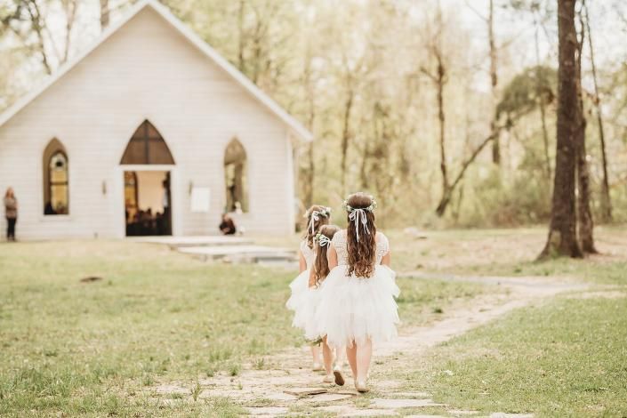Two girls in white dresses walk toward a small chapel on a grassy path.