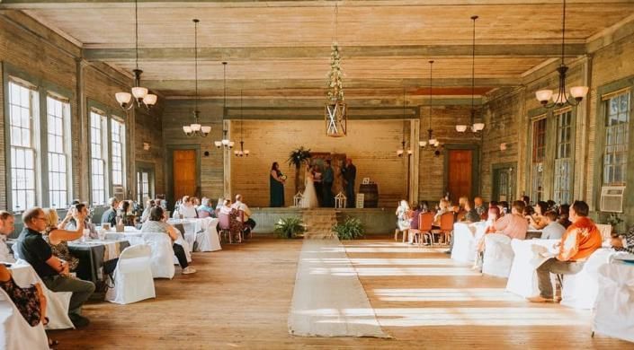 Guests seated at round tables in a sunlit rustic banquet hall facing a decorated stage