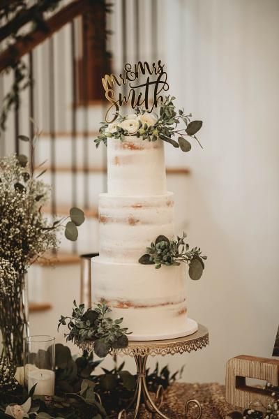 Tiered white wedding cake with greenery and “We Still Do” topper in a rustic indoor setting