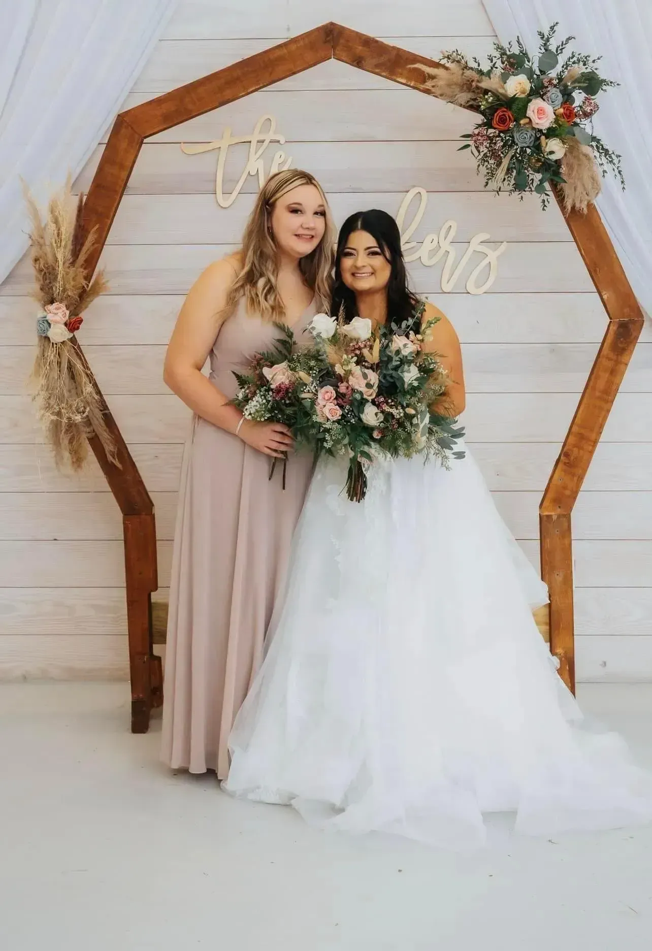 Two brides pose together with bouquets under a wooden arch at a wedding ceremony