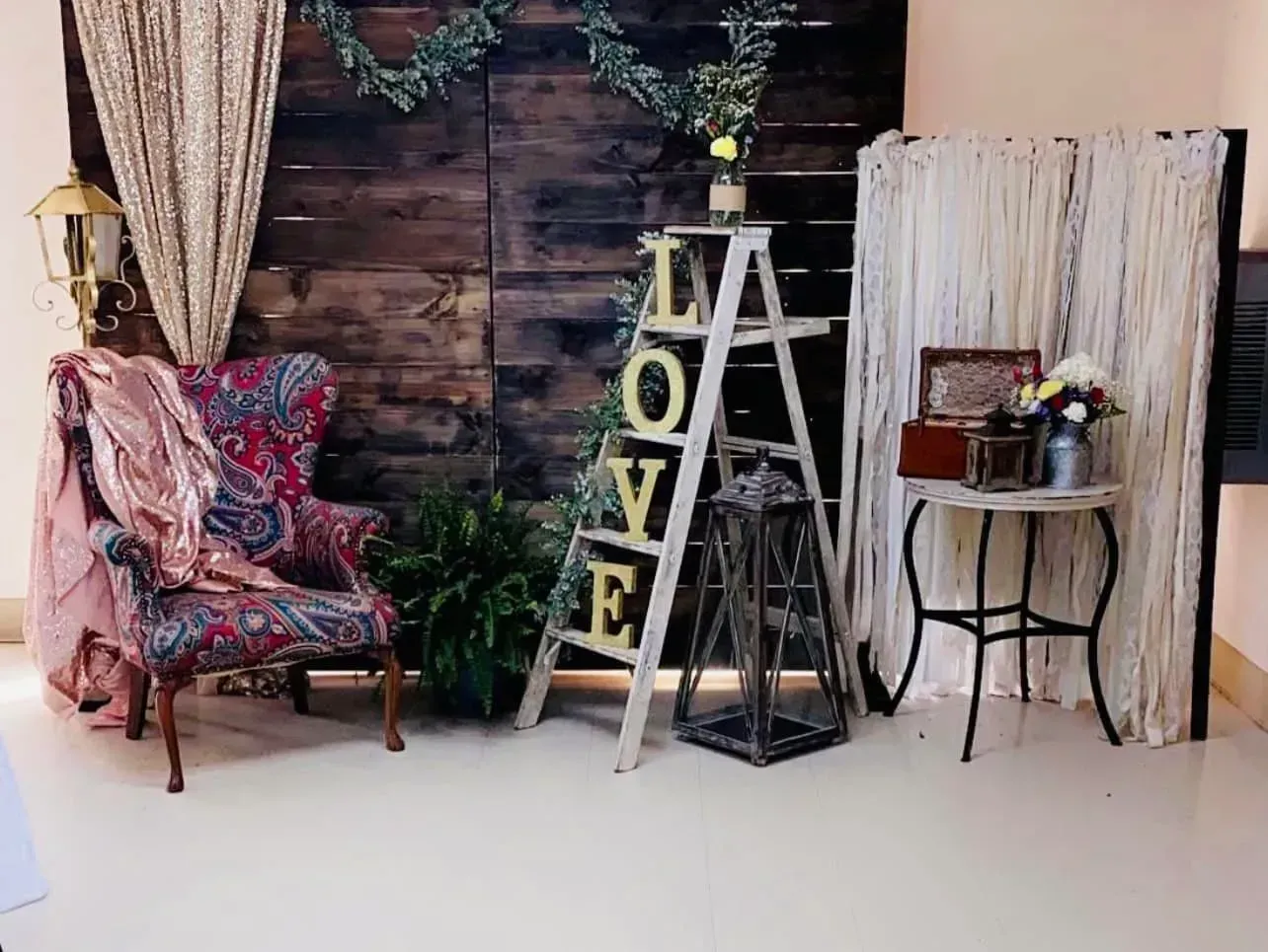 Rustic indoor decor setup with floral chair, ladder shelf, plants, and round side table against a wood backdrop