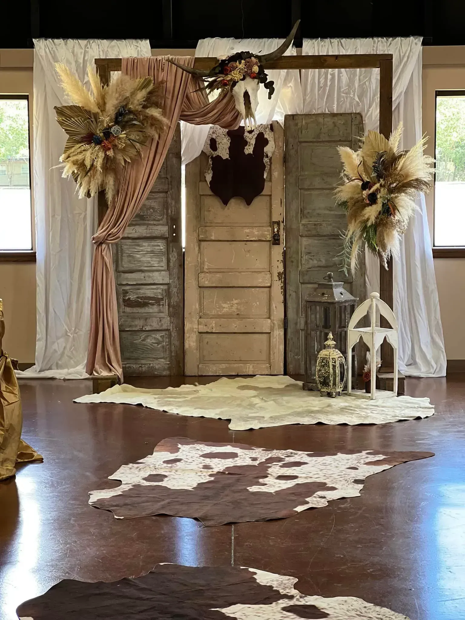 Rustic indoor wedding arch with draped fabric, dried flowers, and wooden doors over white aisle rugs