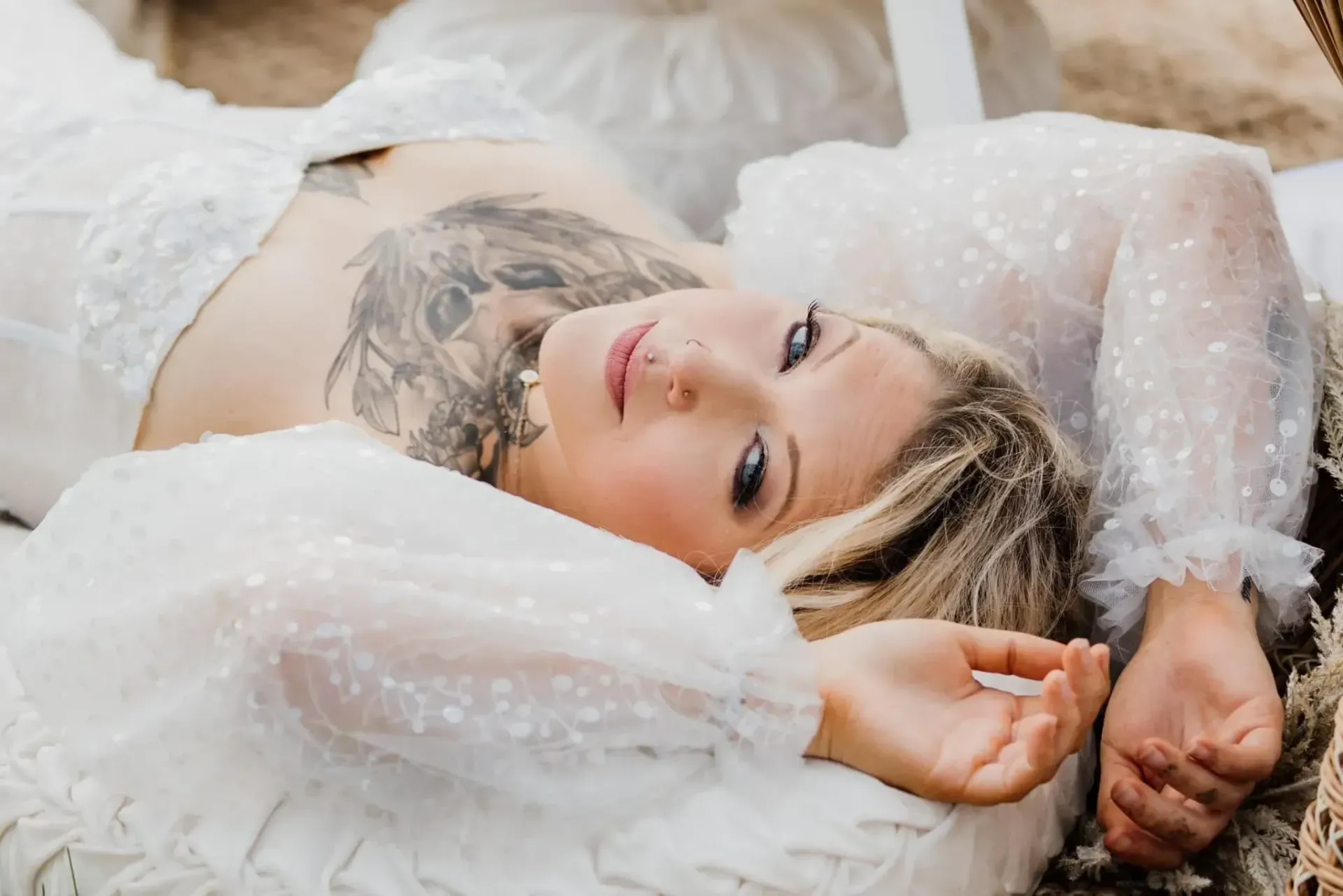 Person lying on white bedding, gazing at the camera, with tattooed chest visible and soft light around them