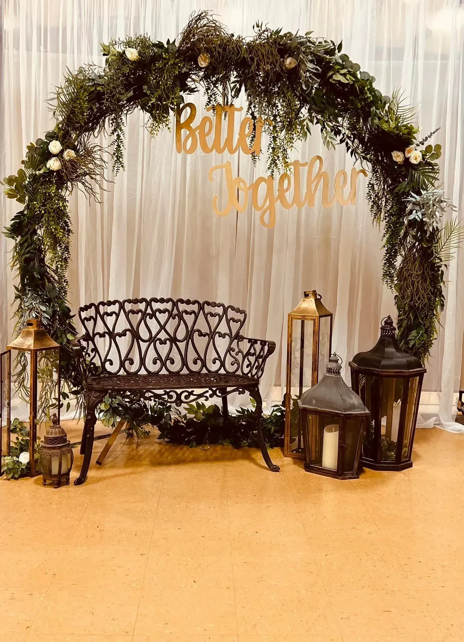 Floral arch over bench and lanterns on a gold carpet, with a sheer backdrop and gold hanging sign.