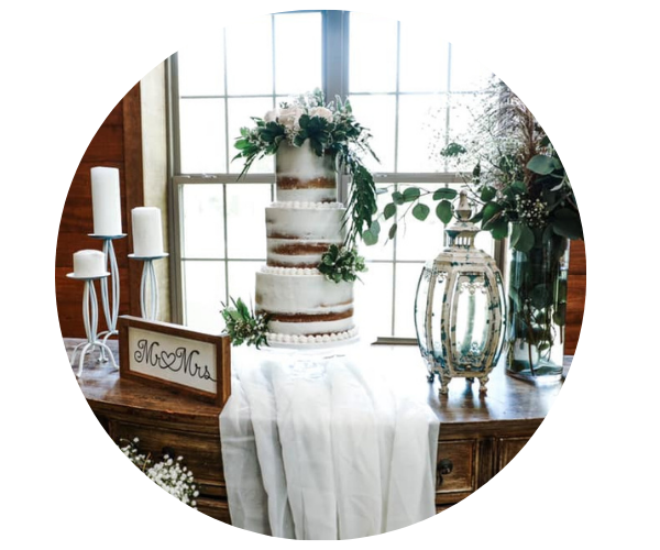 Rustic wedding cake on a draped table with candles, greenery, and glass vases by a bright window