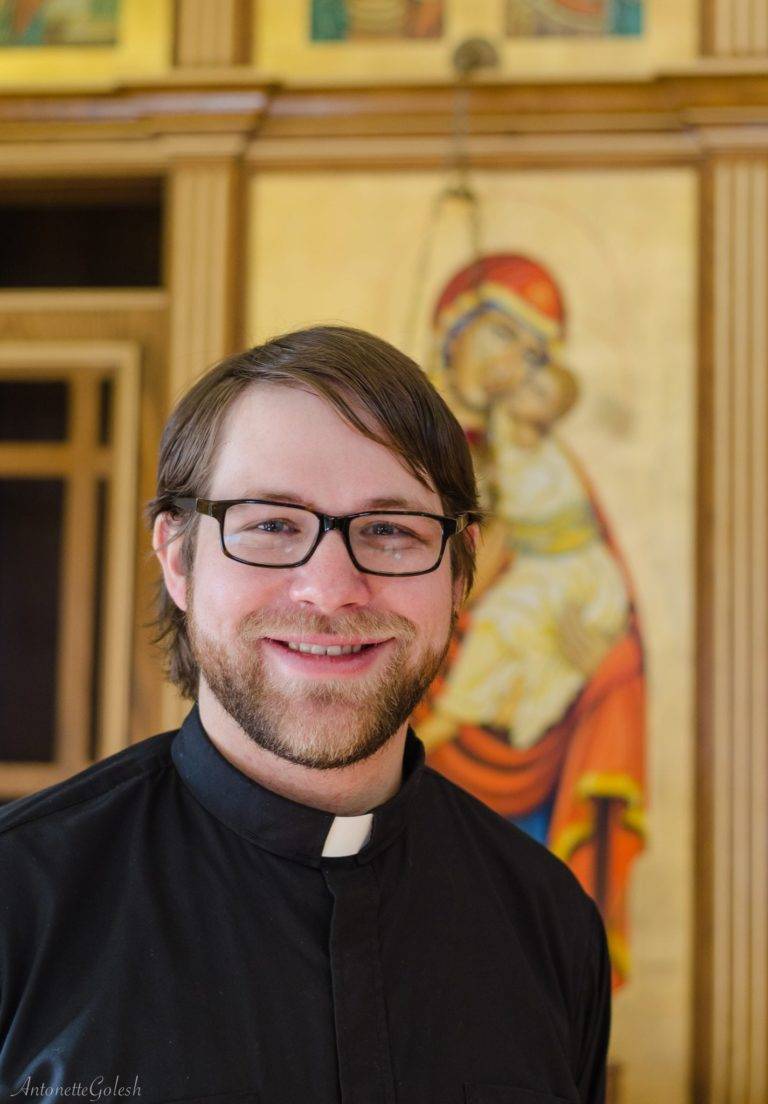 A man with glasses and a beard is smiling in front of a painting of jesus and mary.