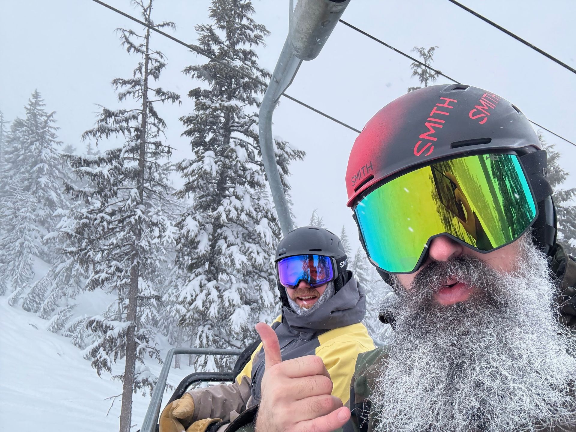 A man with a beard is giving a thumbs up while riding a ski lift.