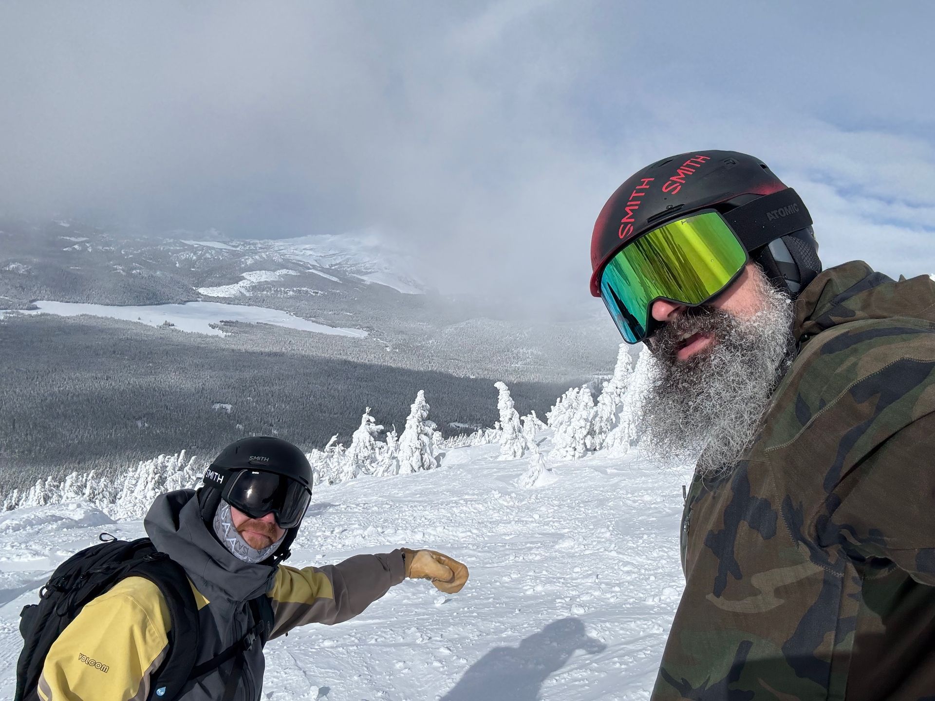 A man with a beard wearing a helmet and goggles is standing next to another man in the snow.