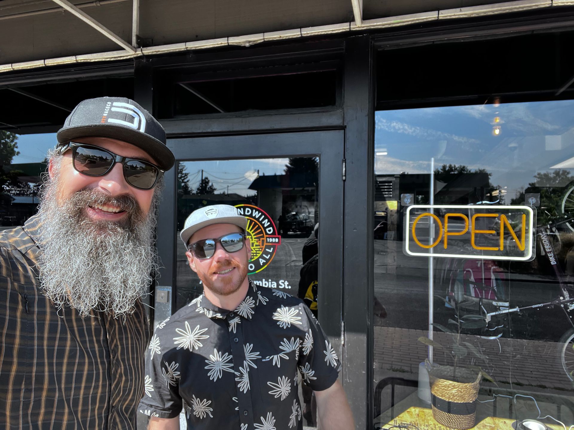 Two men are standing in front of a store with a neon sign that says open.
