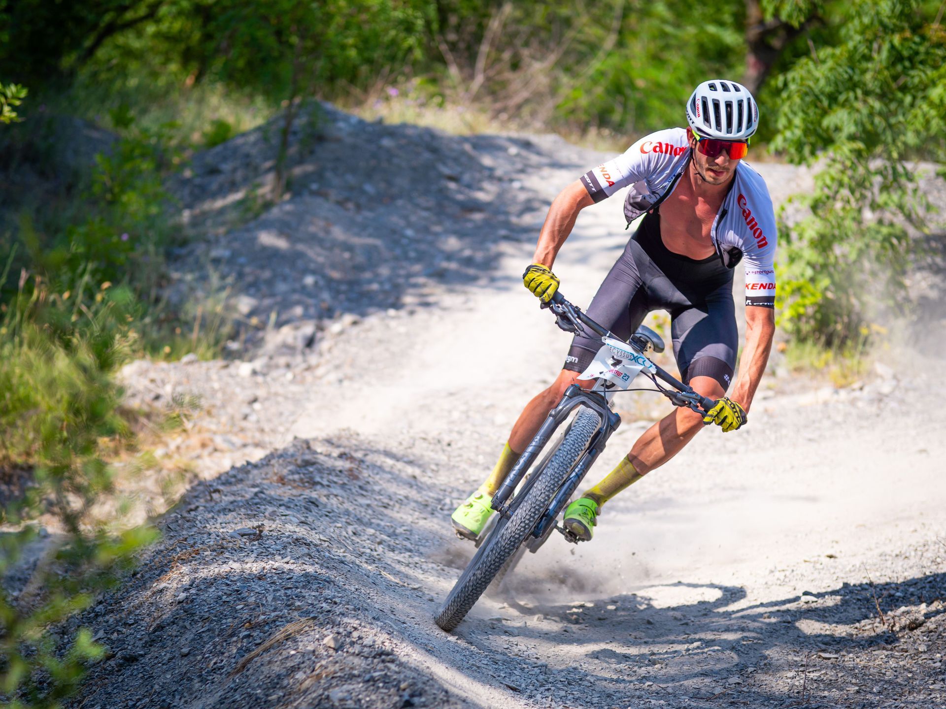 Mountain biker in a helmet and red jersey leans into a turn on a dirt trail, kicking up dust.