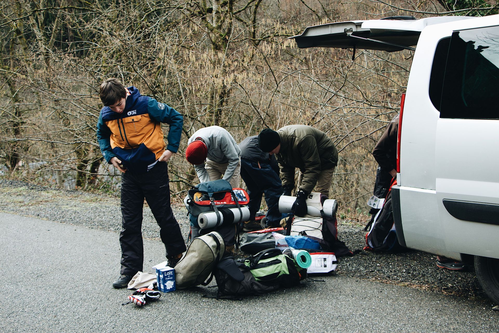 Three cyclists kneel beside fallen bikes and gear next to a white van on a roadside.