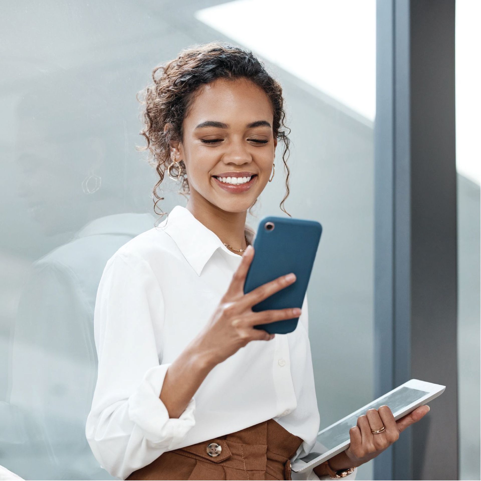 Smiling woman holding a phone and tablet, wearing a white shirt and brown pants, indoors by a window.