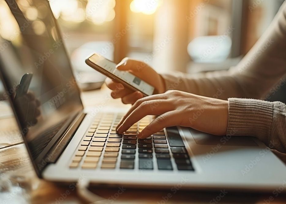 Person typing on laptop, holding phone, possibly multitasking in natural light.