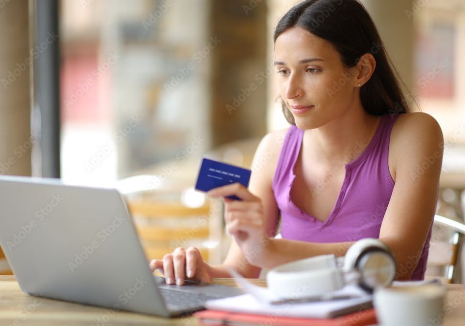 Woman in purple top uses laptop, holds blue credit card at a table.