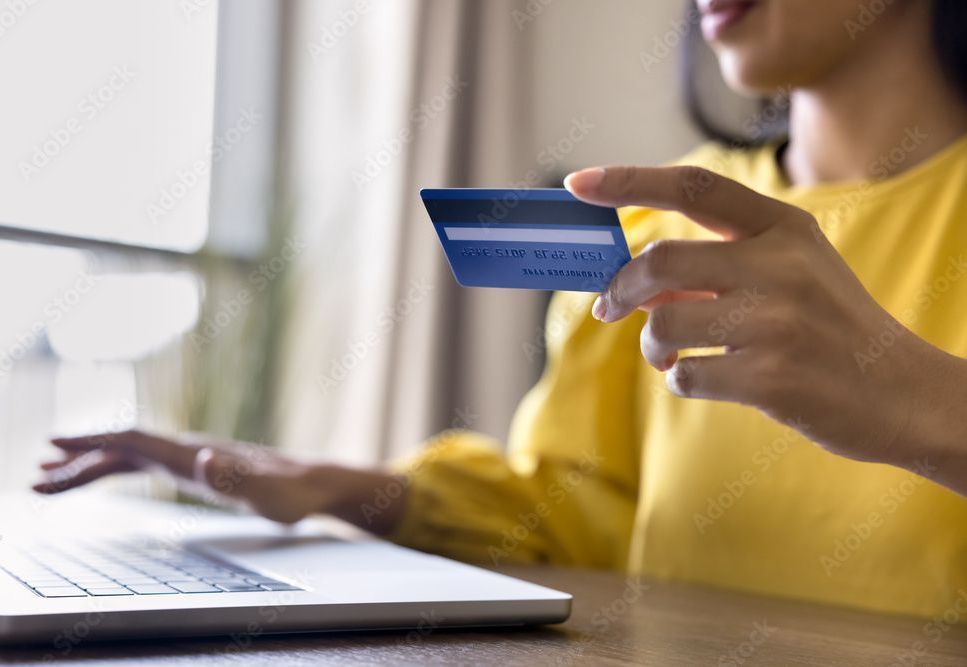Woman holding a blue credit card, entering information on a laptop; yellow shirt, indoor setting.