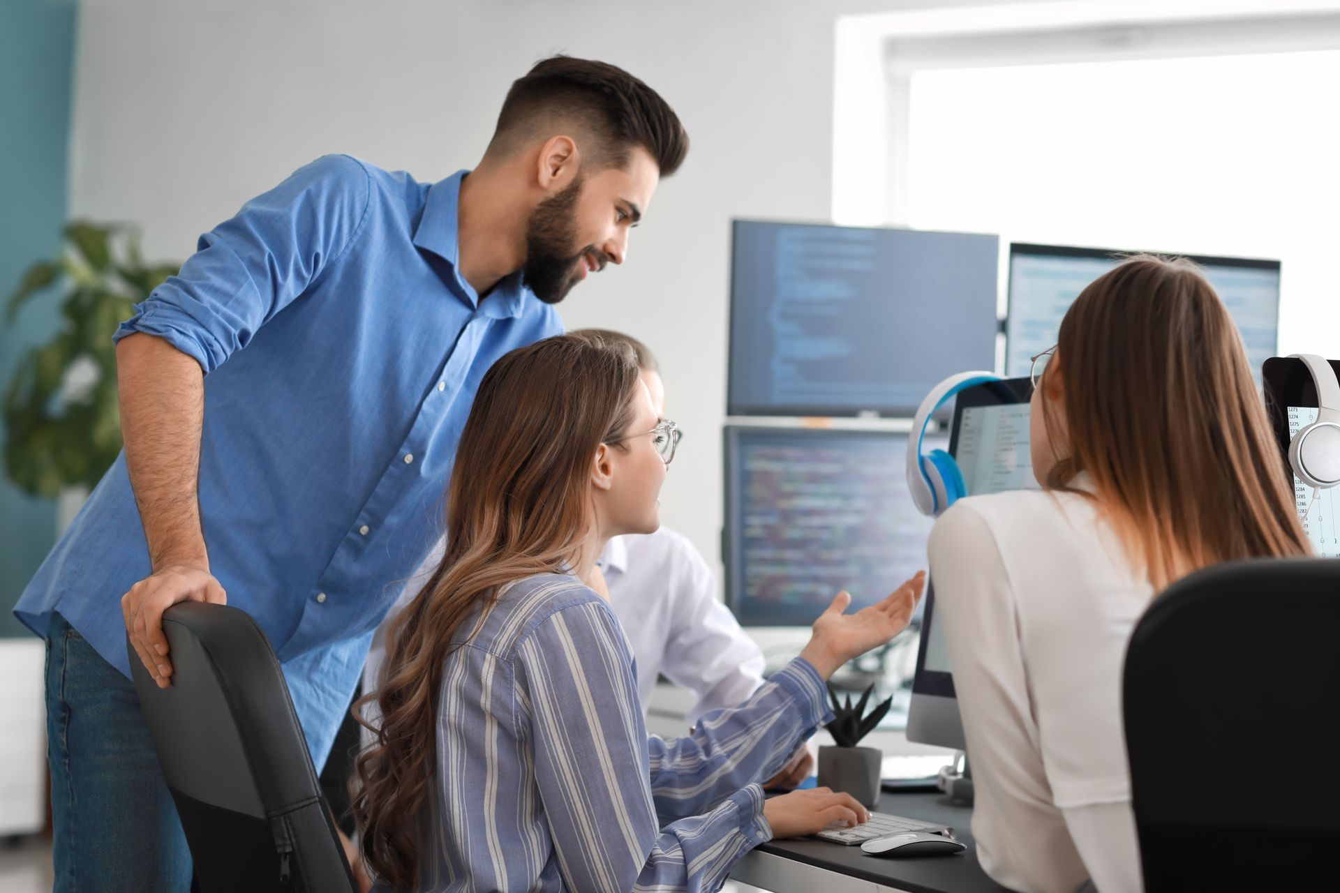 Three people collaborating at computer monitors, possibly coding, in an office setting.