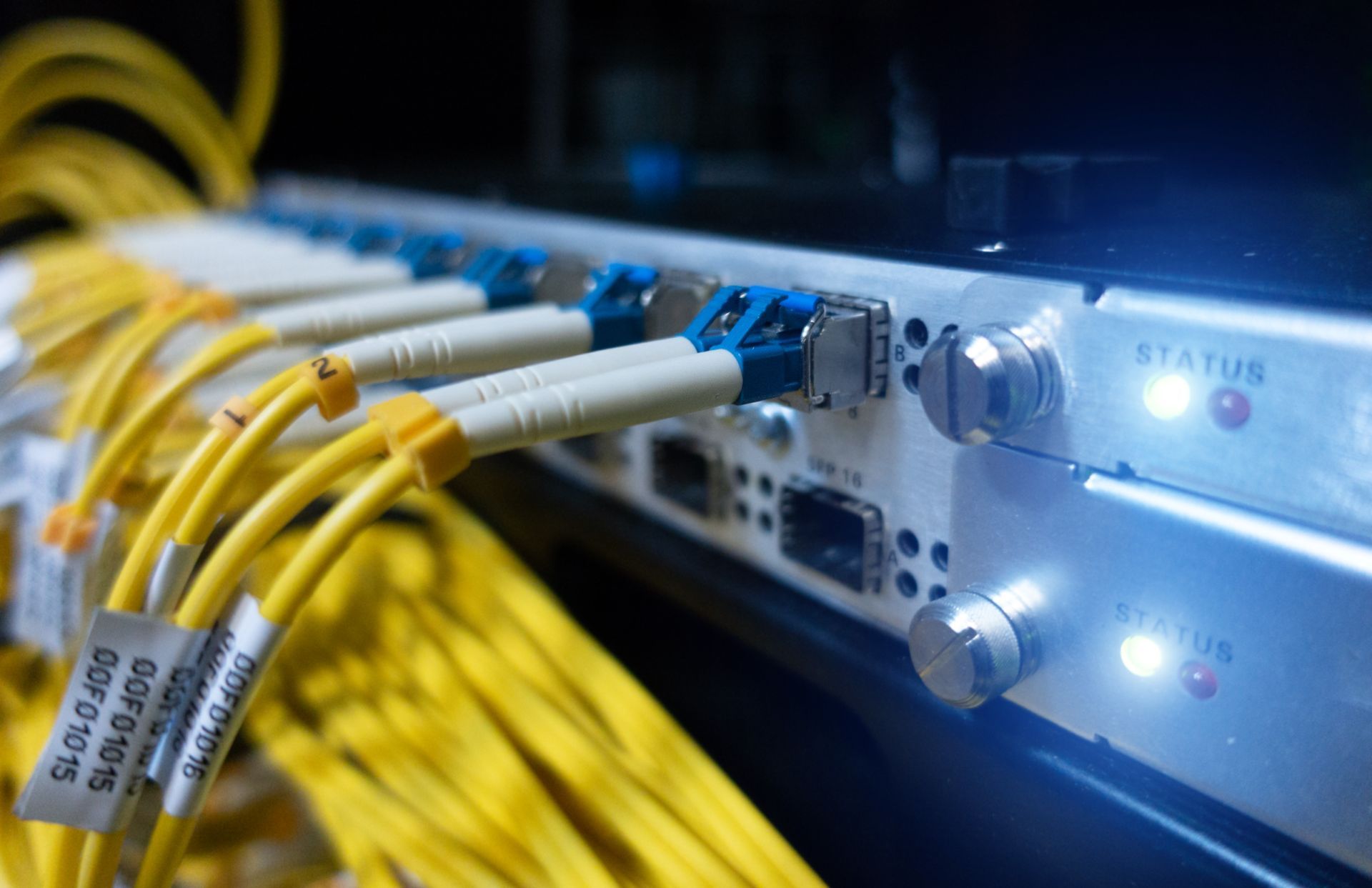 Server rack with yellow fiber optic cables plugged into a white panel. Blue connectors and status lights visible.
