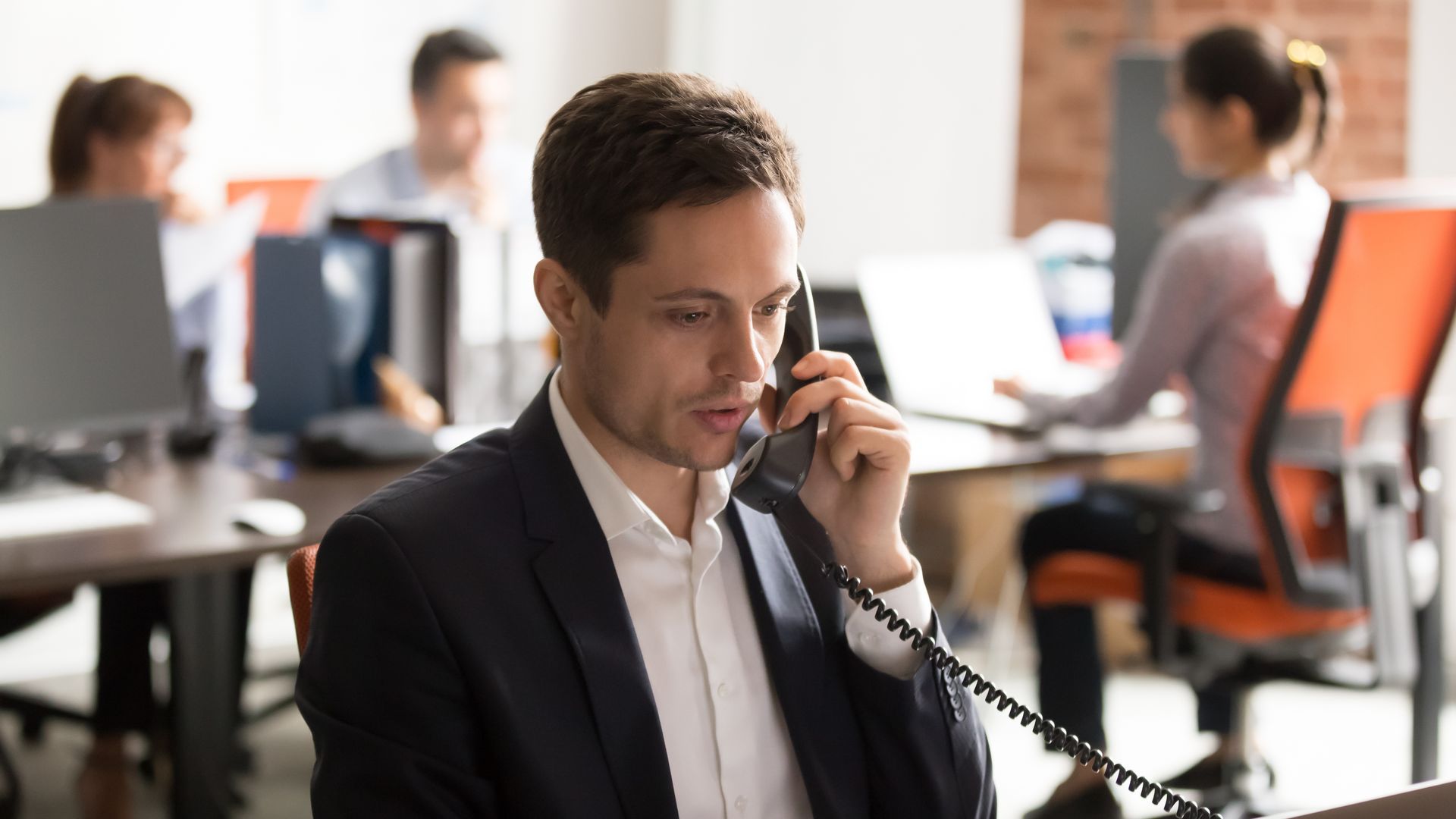 Man in suit on phone in office; coworkers in background.