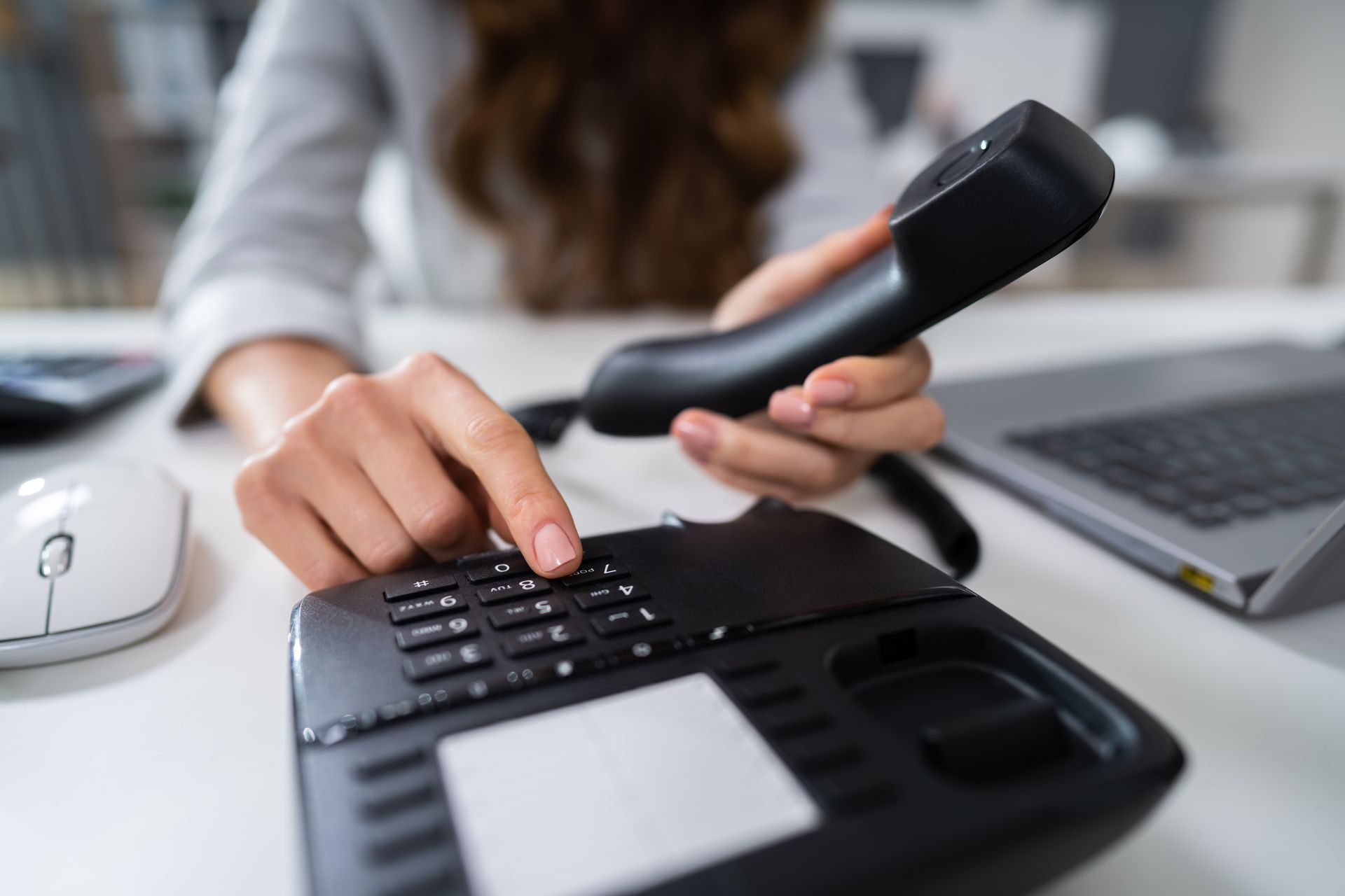 Person dialing a black office phone with a black receiver, desk, and computer.
