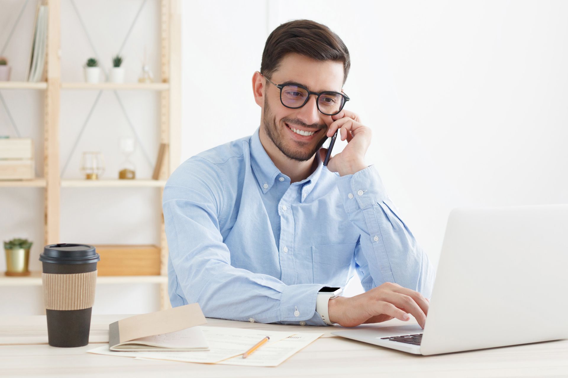 Man in glasses smiling, on a phone call, working on a laptop at a desk with coffee.