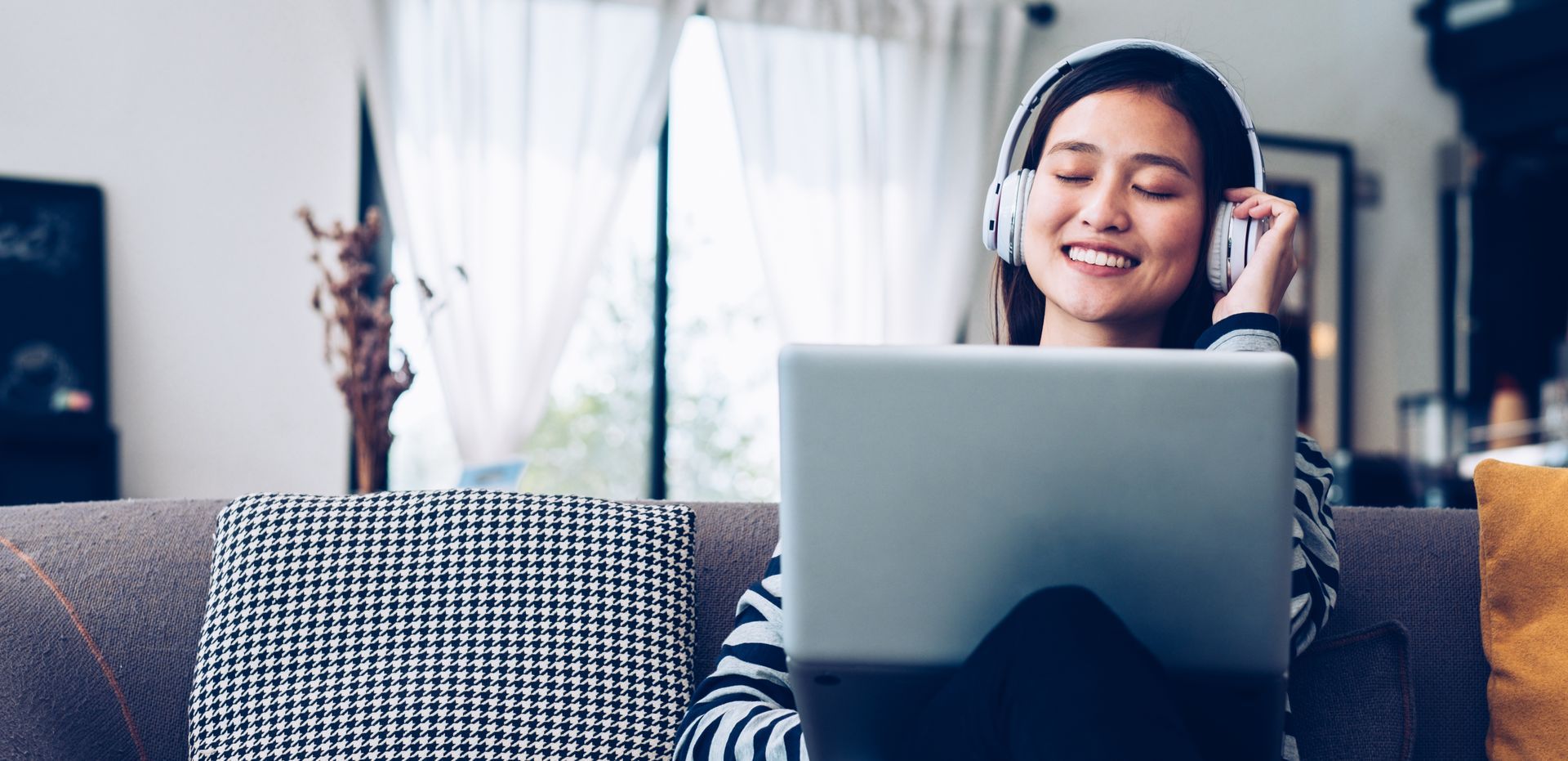 Woman wearing headphones smiles while using a laptop on a couch at home.