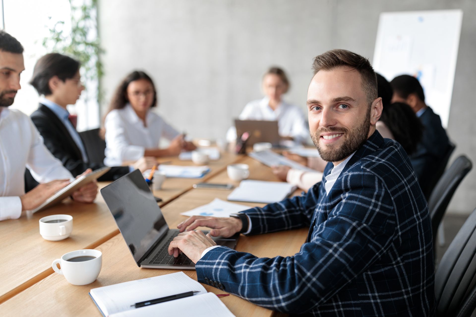 Man in plaid jacket smiles at camera, working on laptop at a conference table with colleagues.