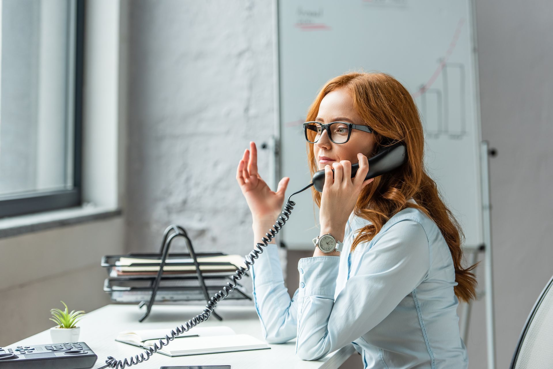 Woman with red hair, glasses, and light blue shirt on the phone at her desk, with a concerned expression.