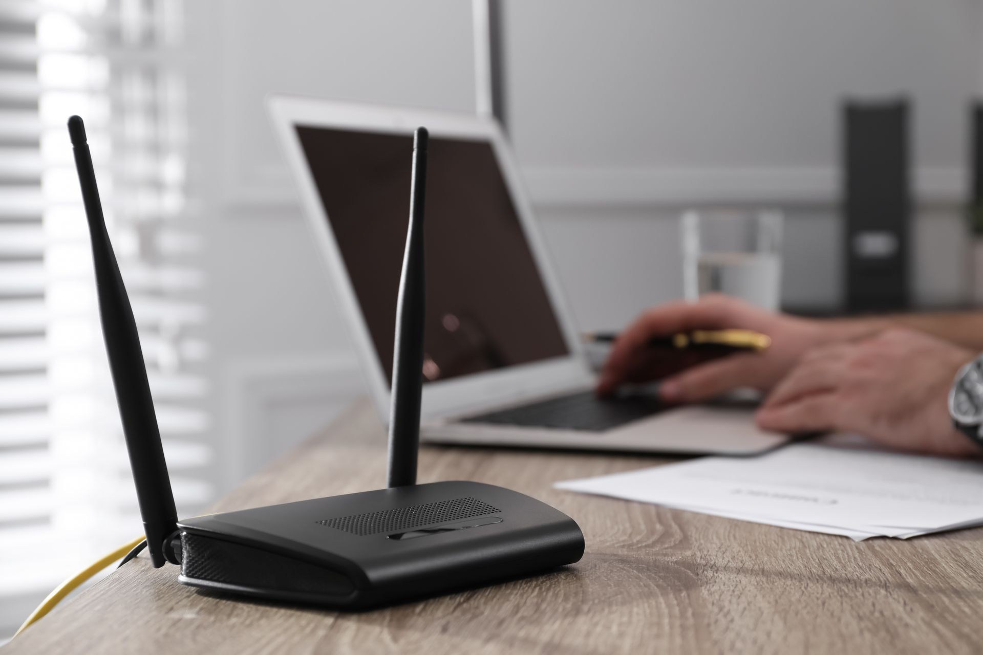Black Wi-Fi router with two antennas on a desk near a laptop and a person typing.