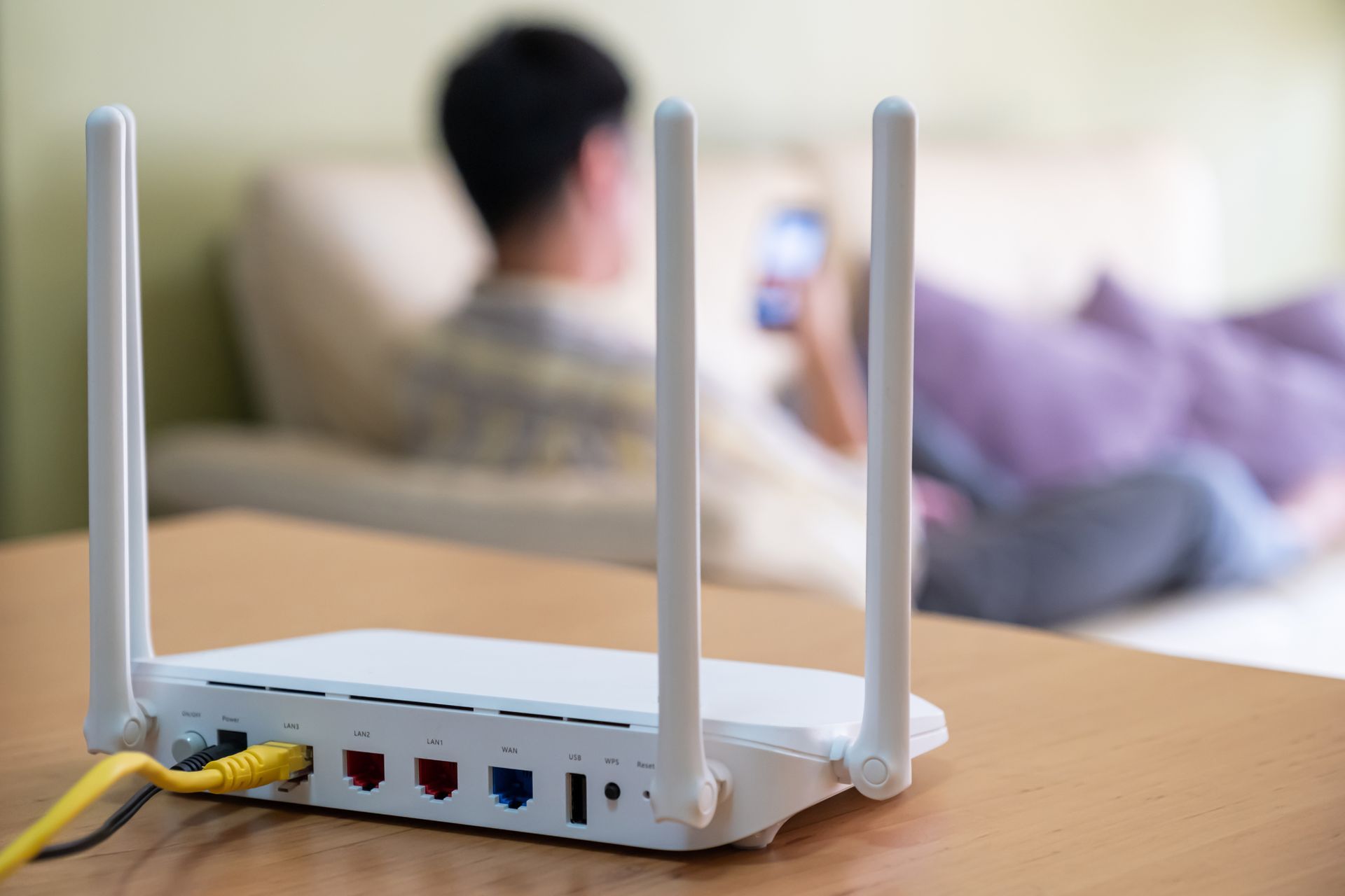 White WiFi router with antennas on a wooden table; person using phone on a couch in the background.