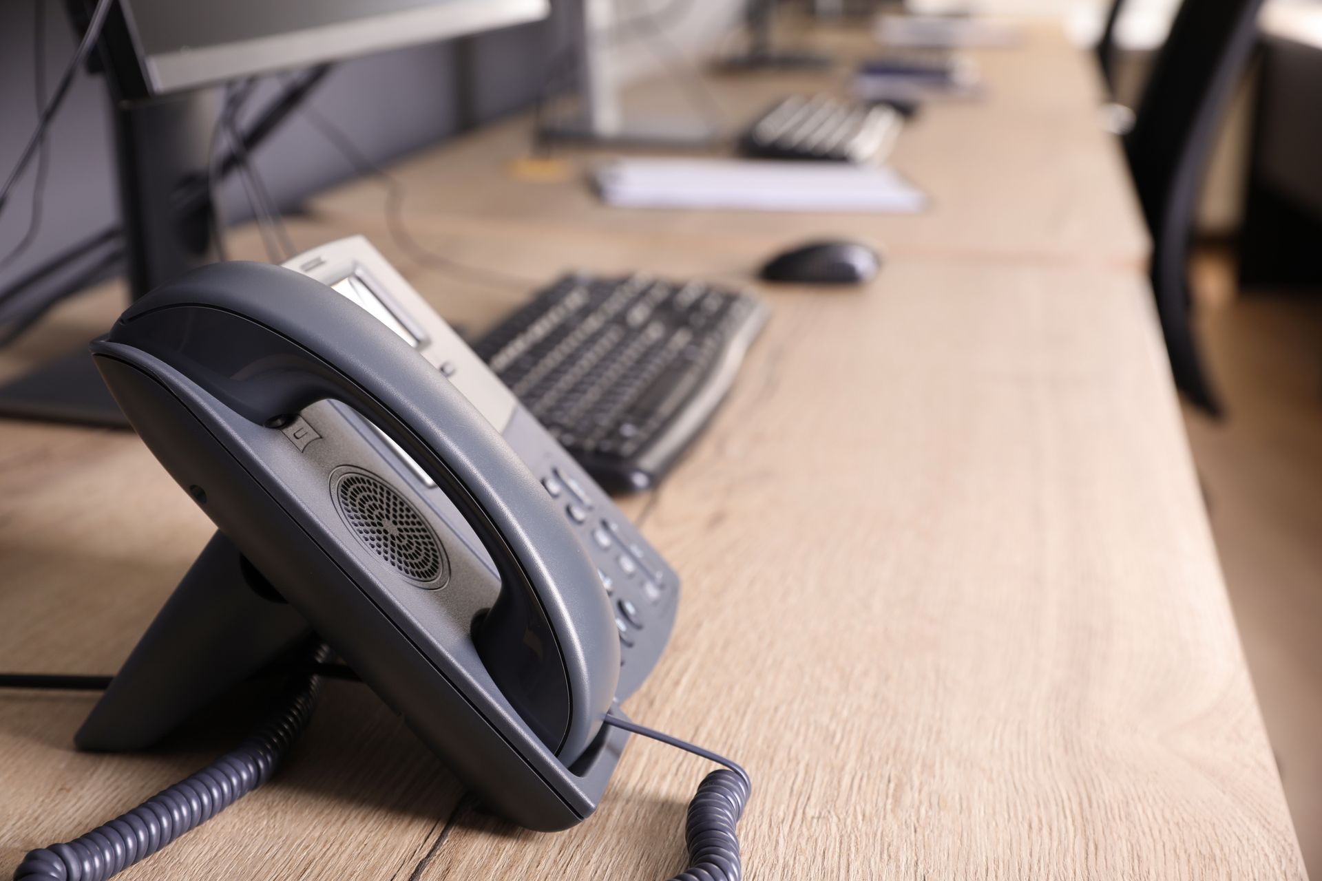 Desk phone and keyboard on a wooden desk, with computer monitors in the background.