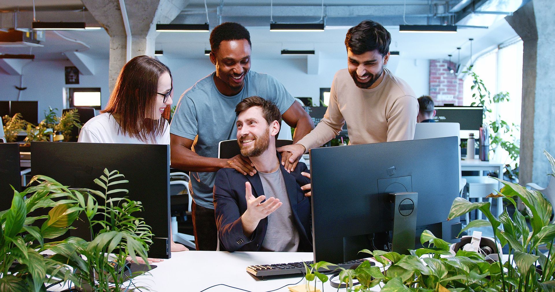 Four coworkers smile, celebrating at a desk, looking at a computer screen in a bright office.