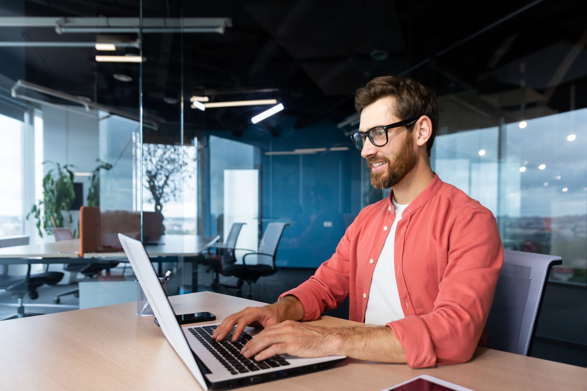 Man with glasses smiles while typing on a laptop in a modern office.