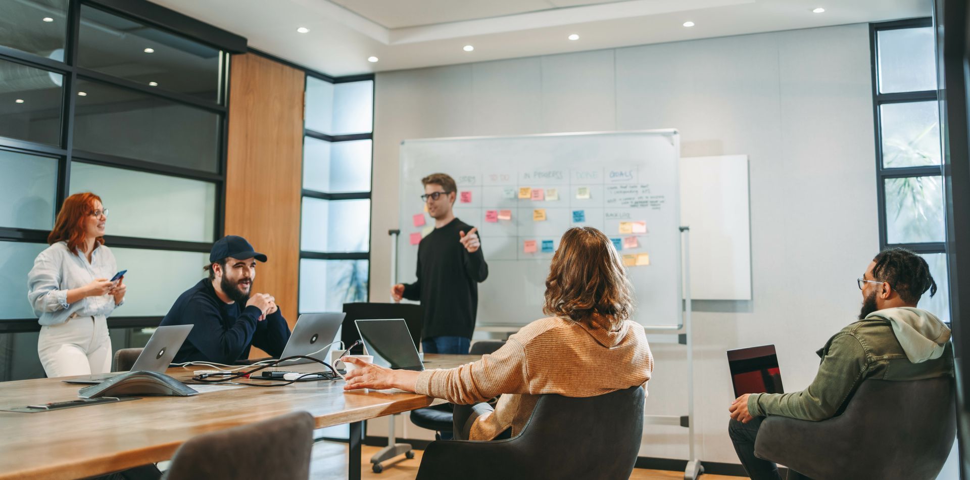 People in a conference room. A man stands by a whiteboard presenting. Others sit at a table with laptops.