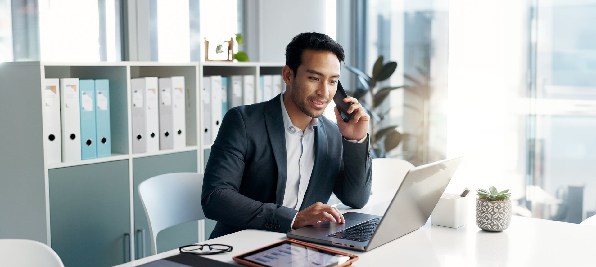 A man in a suit working on a laptop and talking on the phone in an office.