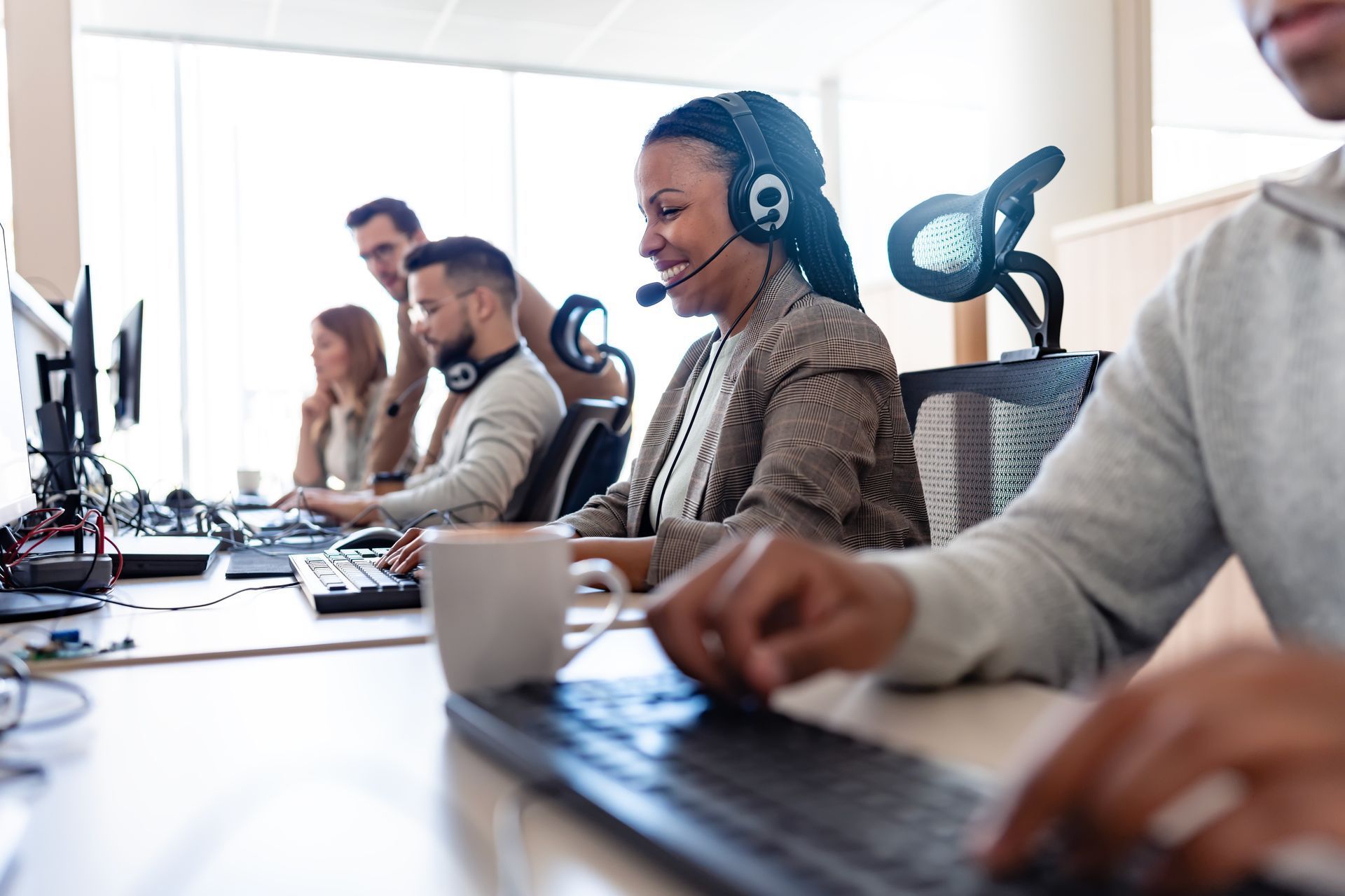Office scene: diverse people working at computers, some wearing headsets, focused, light-filled room.