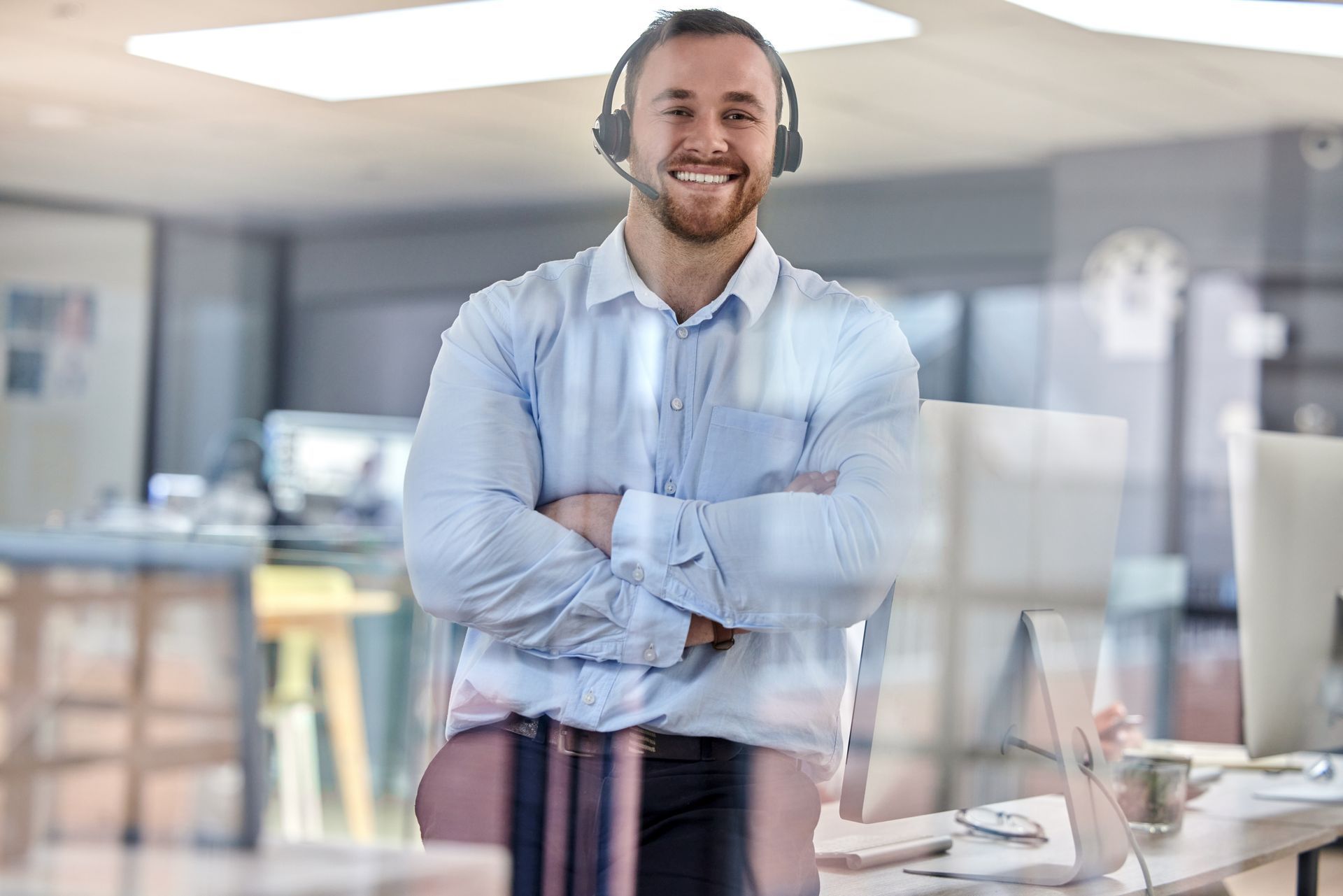 Smiling man with headset, arms crossed, standing in an office.